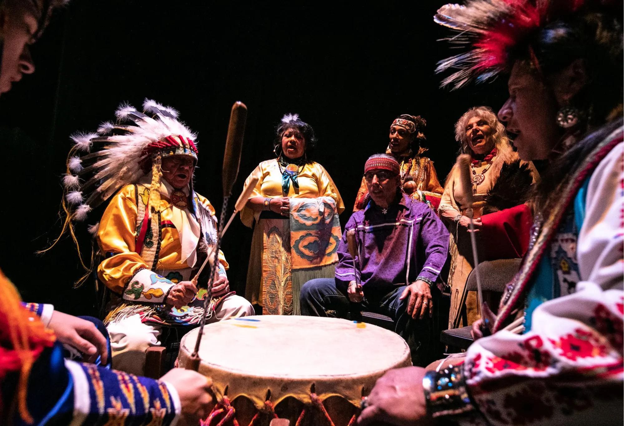 Indigenous performers in various regalia seated around a drum