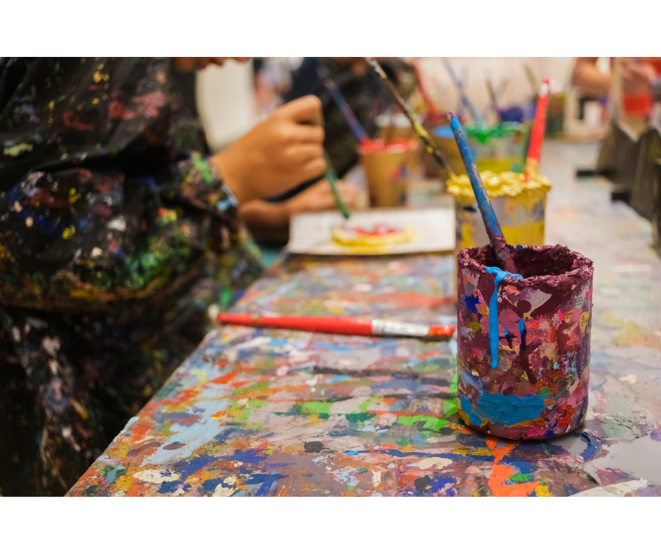 closeup of hands painting canvases with a paint-covered can and paint brushes in the foreground