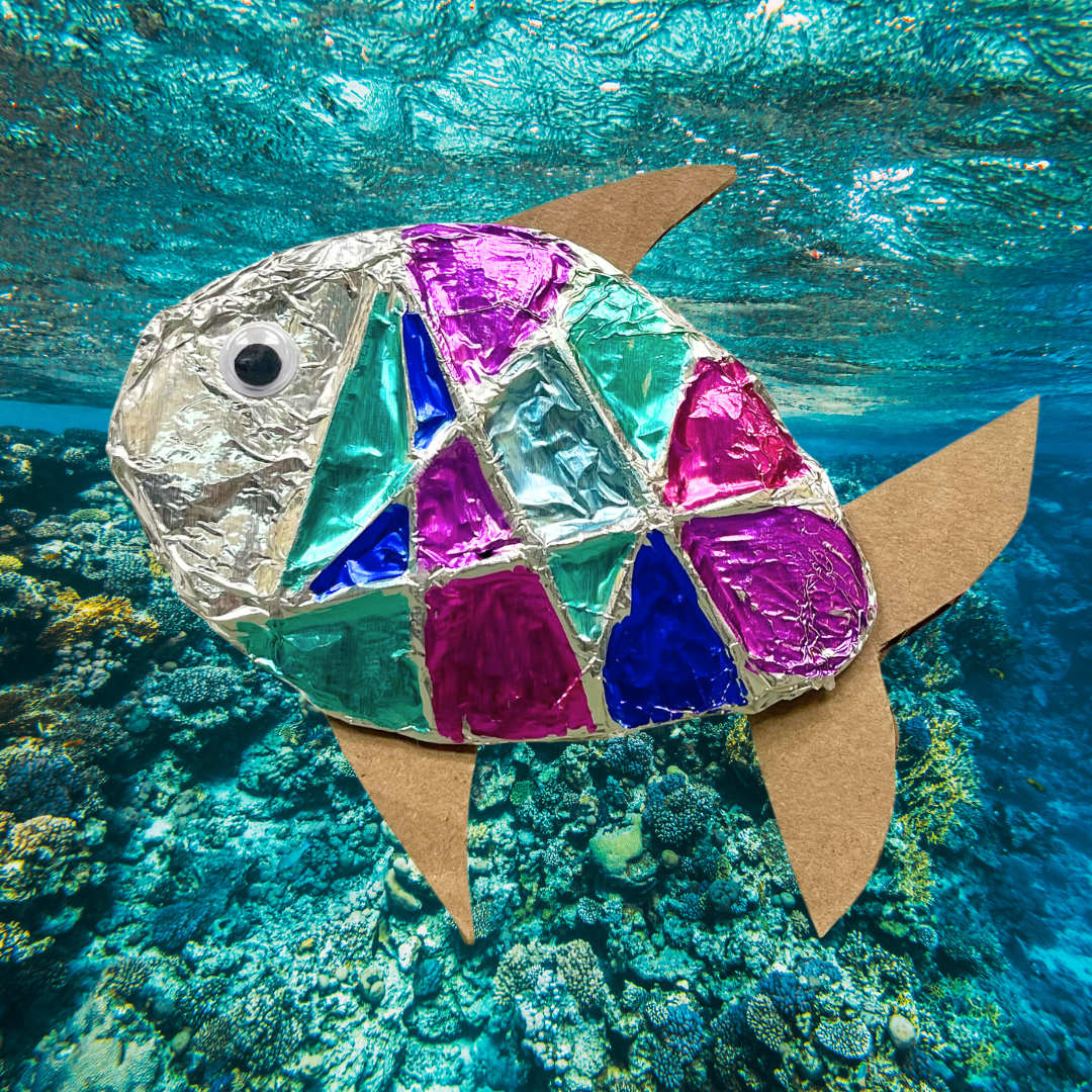 a colorful fish made of tin foil with cardboard fins in front of a photo of a blue reef environment